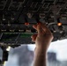 A hand is reaching up to manipulate a switch or control on an aircraft overhead panel in a cockpit. The panel contains numerous switches, toggle controls, indicator lights, and a small green display screen. Several amber/orange warning or status indicator lights are illuminated on the panel.
