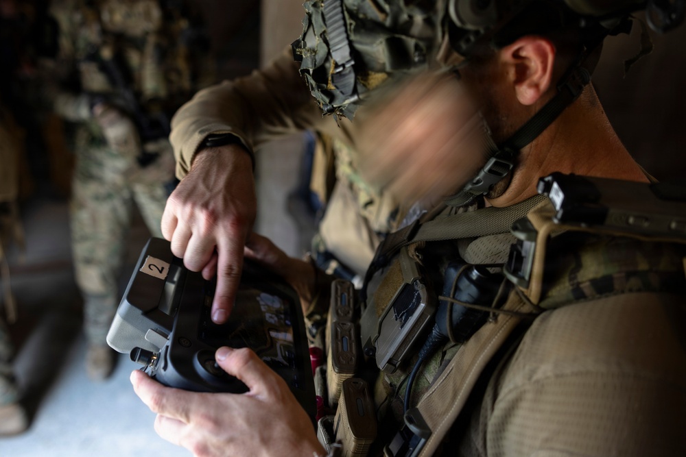 A military personnel in full tactical gear is operating a handheld electronic device, likely a drone controller or tactical tablet. The soldier is wearing camouflage uniform with a tactical vest and a helmet with camouflage netting. A second armed soldier is visible in the blurred background.