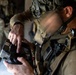 A military personnel in full tactical gear is operating a handheld electronic device, likely a drone controller or tactical tablet. The soldier is wearing camouflage uniform with a tactical vest and a helmet with camouflage netting. A second armed soldier is visible in the blurred background.
