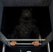 A military personnel wearing digital camouflage (MARPAT pattern consistent with US Marines) and a tactical helmet is ascending a ladder or hatch from below. The perspective is a downward-looking angle from above the opening, showing the soldier gripping metal rungs as they climb upward. The setting appears to be a concrete structure with a hatch or access point.