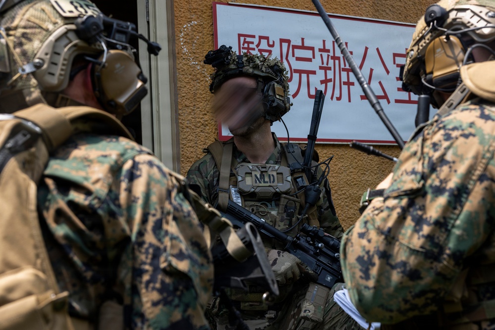 The image shows armed military personnel in full tactical gear positioned near a building with Chinese-language signage. The soldiers are wearing digital camouflage uniforms consistent with U.S. Marine Corps MARPAT pattern, along with advanced communication headsets and tactical vests. The scene appears to depict a military training exercise or operational briefing near a structure with Chinese characters.