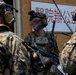 The image shows armed military personnel in full tactical gear positioned near a building with Chinese-language signage. The soldiers are wearing digital camouflage uniforms consistent with U.S. Marine Corps MARPAT pattern, along with advanced communication headsets and tactical vests. The scene appears to depict a military training exercise or operational briefing near a structure with Chinese characters.
