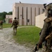 Three heavily equipped military personnel in full tactical gear are advancing along a gravel path toward a tan-colored multi-story training facility complex. The soldiers are wearing camouflage uniforms, tactical helmets with mounted devices, body armor, and carrying weapons. The setting appears to be a military training environment, likely a mock urban operations or MOUT (Military Operations on Urban Terrain) facility.