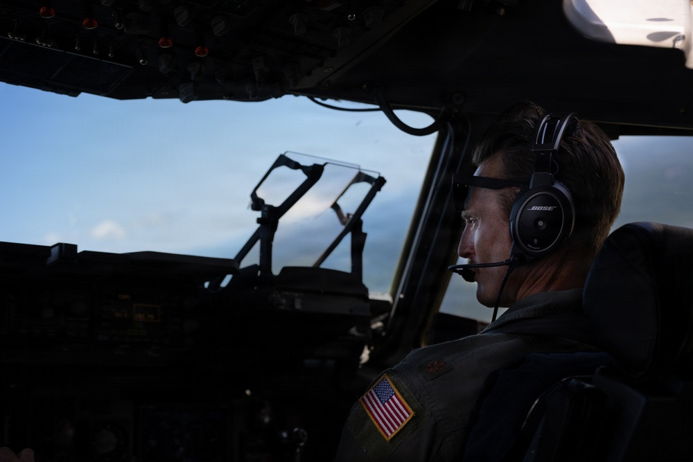 A military pilot is seated in the cockpit of a large aircraft, wearing a Bose aviation headset and a flight suit with a U.S. flag patch on the shoulder. The cockpit is dimly lit with the blue sky visible through the windscreen. The image appears to be taken during flight operations.