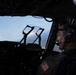 A military pilot is seated in the cockpit of a large aircraft, wearing a Bose aviation headset and a flight suit with a U.S. flag patch on the shoulder. The cockpit is dimly lit with the blue sky visible through the windscreen. The image appears to be taken during flight operations.