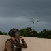 A heavily equipped special operations soldier kneels against a concrete wall while observing a fixed-wing aircraft or drone flying in overcast skies. The soldier is holding a notepad and pen, suggesting active coordination or documentation of the aerial asset. The face has been deliberately blurred, indicating operational security measures.