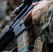A close-up photograph of a military service member holding an assault rifle, likely an M4 carbine or similar AR-platform weapon. The individual is wearing MARPAT (Marine Pattern) digital camouflage, indicating affiliation with the United States Marine Corps. A tan/FDE Magpul PMAG magazine is inserted into the weapon.