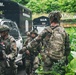 The image shows a group of armed military or law enforcement personnel in camouflage uniforms operating near vehicles marked 'U.C.M.' in a tropical, jungle-like environment. At least three individuals are visible, equipped with tactical gear, helmets, and firearms. The scene appears to depict a coordinated tactical operation or training exercise.