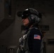 A female military aviator or aircrew member is shown in profile wearing full flight gear including a helmet with visor and communications equipment. She is wearing a flight suit with a US flag patch on the left shoulder and a tactical vest with harness equipment. The setting appears to be inside a military aircraft hangar or similar facility.