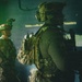 Two heavily equipped military personnel are positioned inside what appears to be a damaged or smoke-filled building. The foreground soldier is wearing full tactical gear including a ballistic helmet with night-vision mount, multicam plate carrier, and backpack. The background soldier displays a patch consistent with the Peruvian flag on their left arm.