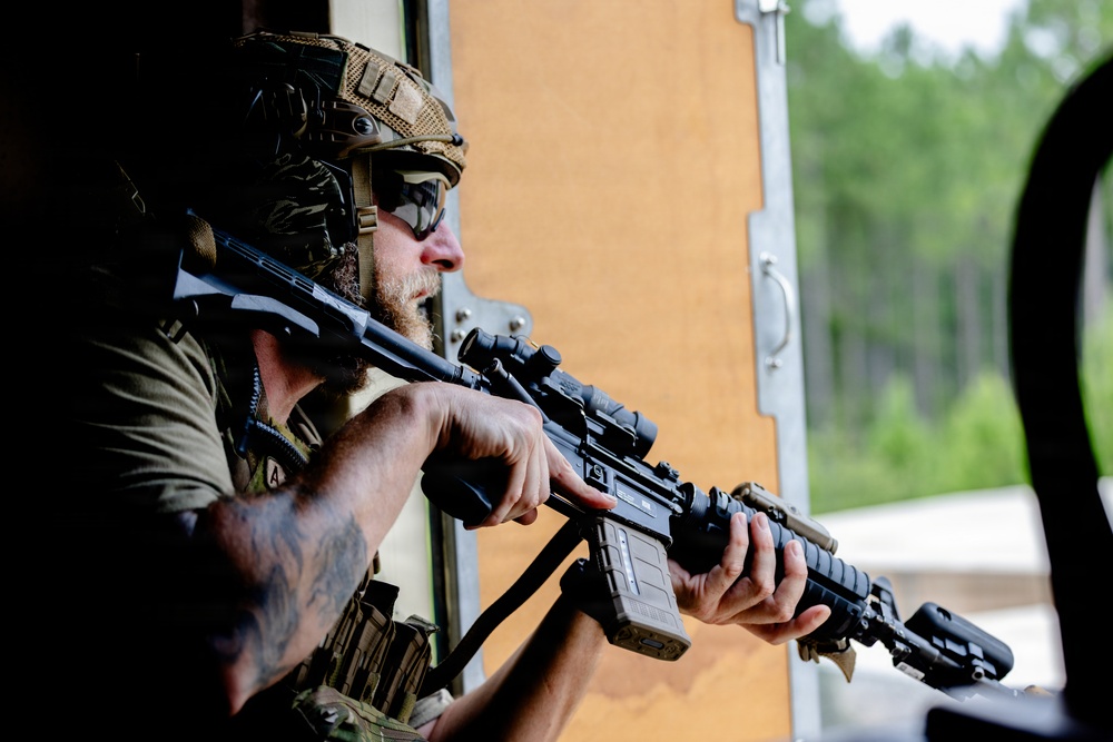 A heavily equipped tactical operator is positioned at an opening, holding an AR-platform rifle with an optic scope and extended magazine. The individual is wearing a tactical helmet, protective eyewear, plate carrier vest, and has visible tattoos on their arms. The setting appears to be a training facility or range environment based on the constructed structure and wooded background.