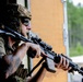 A heavily equipped tactical operator is positioned at an opening, holding an AR-platform rifle with an optic scope and extended magazine. The individual is wearing a tactical helmet, protective eyewear, plate carrier vest, and has visible tattoos on their arms. The setting appears to be a training facility or range environment based on the constructed structure and wooded background.