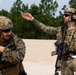 Two armed military personnel in full combat gear are engaged in what appears to be a briefing or instruction session in an outdoor sandy training area. The soldier on the right, wearing OCP (Operational Camouflage Pattern) uniform, is gesturing with one hand while holding papers, appearing to instruct the other. The soldier on the left wears USMC MARPAT digital camouflage and has a helmet label reading 'NOT A JTAC'.