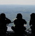 Three military personnel are silhouetted against an aerial landscape, seated on what appears to be the open rear ramp of a military transport aircraft in flight. All three individuals are wearing flight helmets with communication headsets and appear to be observing or photographing the terrain below. The landscape visible beneath them includes forested hills, a body of water, and what appears to be a suburban or semi-rural area.