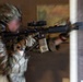 A soldier in full multicam camouflage gear is conducting a close-quarters building clearance drill, aiming an assault rifle toward a doorway or wall corner. The image is intentionally motion-blurred, suggesting dynamic movement during a training exercise. A yellow training/safety marker is visible on the muzzle of the weapon, indicating this is a training scenario rather than live combat.