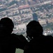 Two silhouetted individuals wearing military-style helmets and gear are viewed from behind, looking out over a densely populated urban area from a significant elevation, likely from an open aircraft door or ramp. The foreground figures are in deep shadow while the cityscape background is brightly lit, creating a strong contrast. One figure appears to have a flag patch visible on their arm.