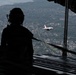 The image is taken from the open rear cargo ramp of a large military transport aircraft, likely a C-17 Globemaster III, showing two silhouetted crew members observing a U.S. Air Force Thunderbirds F-16 jet flying in close formation alongside. The Thunderbird aircraft is clearly visible in its distinctive white and red livery against a backdrop of a suburban landscape far below. The perspective creates a dramatic contrast between the dark interior of the cargo bay and the bright exterior aerial view.