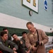 An instructor in a tan/khaki flight suit is demonstrating survival equipment to a group of military personnel seated on bleachers in what appears to be an indoor swimming pool facility. The students, wearing olive drab flight suits, are attentively watching the demonstration. Orange immersion/survival suits and other equipment are visible on the bench nearby.