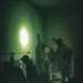 A low-light interior scene showing what appears to be a law enforcement or military operation inside a deteriorated building. A uniformed, helmeted individual with tactical gear is standing next to a civilian whose hands appear to be restrained behind their back. The scene is illuminated by a single bright light source casting strong shadows on the damaged walls.