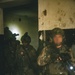 The image shows a group of heavily armed and equipped military or special operations personnel conducting what appears to be a building clearance or close-quarters operation in a darkened, deteriorated structure. One operator in the foreground has their face deliberately blurred, suggesting operational security concerns. At least one patch visible on a uniform appears to be a Peruvian flag, indicating possible Peruvian military or special forces involvement.