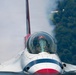 A close-up frontal view of a U.S. Air Force Thunderbirds F-16 Fighting Falcon jet aircraft. The image captures the cockpit canopy with a pilot visible inside wearing a red USAF helmet. The aircraft displays the distinctive Thunderbirds red, white, and blue livery with smoke visible in the background.