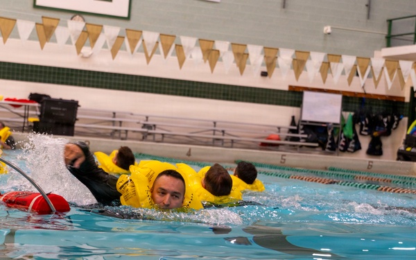 Multiple individuals wearing bright yellow inflatable survival suits are swimming in an indoor pool, appearing to conduct water survival or emergency training. The participants are fully clothed in what appear to be immersion/anti-exposure suits, suggesting military or aviation water survival training. The setting is a standard indoor competitive swimming pool with lane markers and pennant decorations.