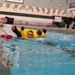 Multiple individuals wearing bright yellow inflatable survival suits are swimming in an indoor pool, appearing to conduct water survival or emergency training. The participants are fully clothed in what appear to be immersion/anti-exposure suits, suggesting military or aviation water survival training. The setting is a standard indoor competitive swimming pool with lane markers and pennant decorations.
