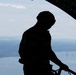 A silhouetted figure wearing a helmet and tactical gear stands at the open ramp or door of a military aircraft in flight. The person appears to be a crew member or loadmaster holding onto a railing while looking downward. In the background, a large body of water, a suspension bridge, and a distant cityscape are visible from altitude.