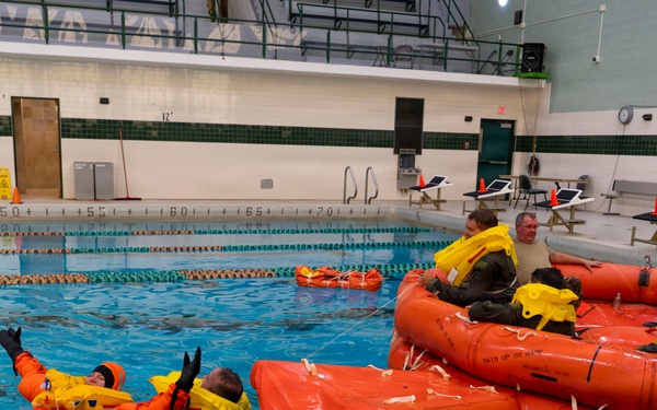 A water survival training exercise is taking place in an indoor swimming pool facility. Participants wearing orange immersion suits and yellow life vests are practicing emergency water survival techniques, including use of inflatable orange life rafts. One individual is floating in the water while others are seated in or around the deployed life rafts.