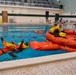 A water survival training exercise is taking place in an indoor swimming pool facility. Participants wearing orange immersion suits and yellow life vests are practicing emergency water survival techniques, including use of inflatable orange life rafts. One individual is floating in the water while others are seated in or around the deployed life rafts.