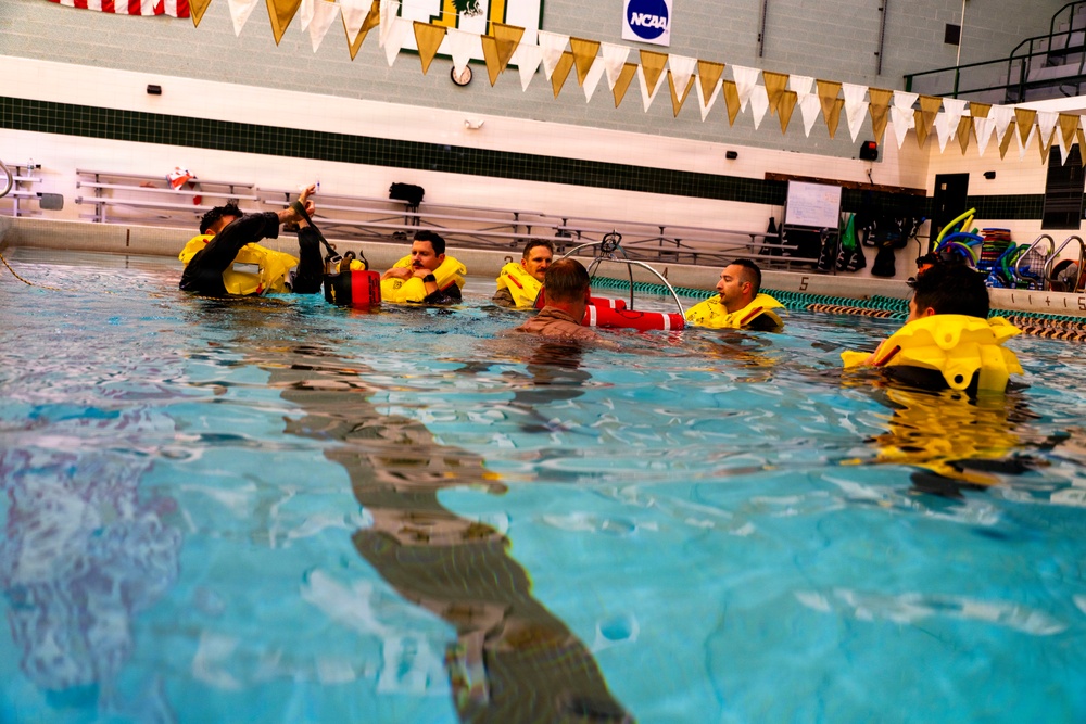 A group of individuals are participating in what appears to be a water survival or rescue training exercise in an indoor swimming pool. Several participants are wearing yellow inflatable life vests/survival suits and are gathered in the pool around an instructor. The setting is an NCAA-affiliated aquatic facility, identifiable by the NCAA banner and pennant decorations on the walls.