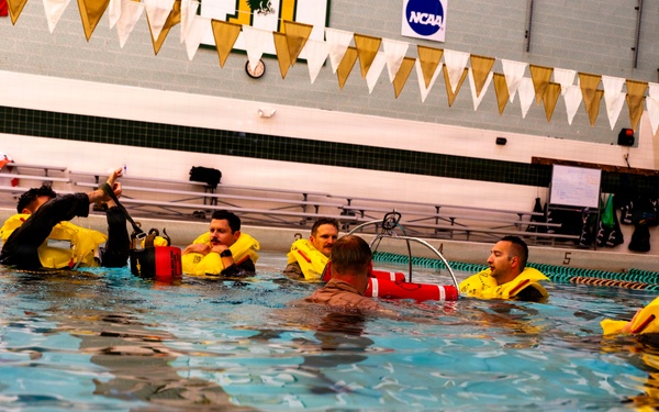 A group of individuals are participating in what appears to be a water survival or rescue training exercise in an indoor swimming pool. Several participants are wearing yellow inflatable life vests/survival suits and are gathered in the pool around an instructor. The setting is an NCAA-affiliated aquatic facility, identifiable by the NCAA banner and pennant decorations on the walls.