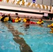 A group of individuals are participating in what appears to be a water survival or rescue training exercise in an indoor swimming pool. Several participants are wearing yellow inflatable life vests/survival suits and are gathered in the pool around an instructor. The setting is an NCAA-affiliated aquatic facility, identifiable by the NCAA banner and pennant decorations on the walls.