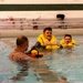 Three individuals are in an indoor swimming pool during what appears to be a water survival or safety training exercise. Two men wearing inflated yellow life vests are in the water facing a third person whose back is to the camera. The man in the center is giving a thumbs-up gesture, suggesting successful completion of a task or positive acknowledgment.