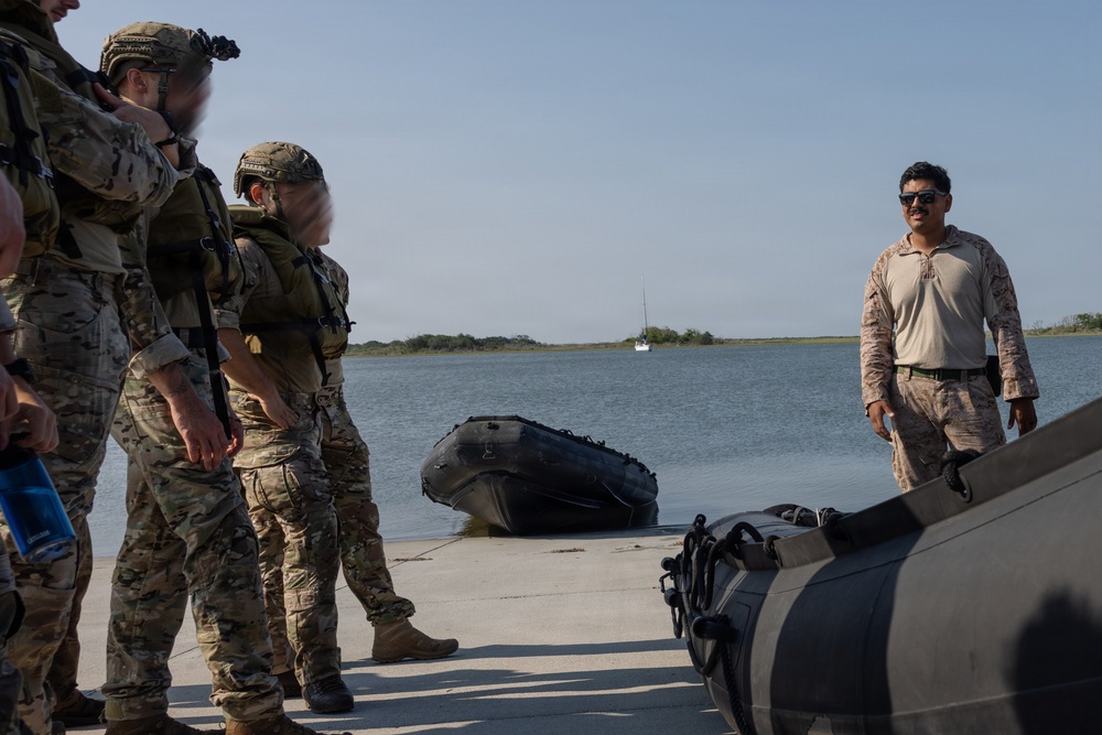 A group of military personnel in tactical gear are gathered at a waterfront location, appearing to receive a briefing or instruction near an inflatable rubber raiding craft (CRRC). One individual in desert camouflage pattern (possibly a different unit or nationality) stands facing the group, suggesting an instructional or coordination role. The setting appears to be a calm inland waterway or bay with a sailboat visible in the background.