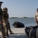 A group of military personnel in tactical gear are gathered at a waterfront location, appearing to receive a briefing or instruction near an inflatable rubber raiding craft (CRRC). One individual in desert camouflage pattern (possibly a different unit or nationality) stands facing the group, suggesting an instructional or coordination role. The setting appears to be a calm inland waterway or bay with a sailboat visible in the background.
