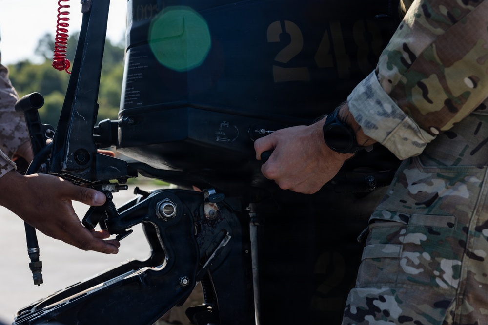 Two military personnel in different camouflage uniforms are working on what appears to be an outboard boat motor marked with the number '24'. One individual is wearing OCP (Operational Camouflage Pattern) uniform while the other appears to be in MARPAT or similar pattern. They are performing maintenance or assembly work on the motor using hand tools.
