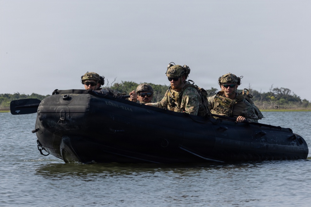 Four military personnel in full combat gear are traveling in a black inflatable rubber raiding craft (CRRC) across a calm body of water. The operators are wearing camouflage uniforms, tactical helmets with communications equipment, body armor, and sunglasses. Vegetation and a shoreline are visible in the background, suggesting a coastal or estuarine environment.