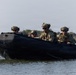 Four military personnel in full combat gear are traveling in a black inflatable rubber raiding craft (CRRC) across a calm body of water. The operators are wearing camouflage uniforms, tactical helmets with communications equipment, body armor, and sunglasses. Vegetation and a shoreline are visible in the background, suggesting a coastal or estuarine environment.