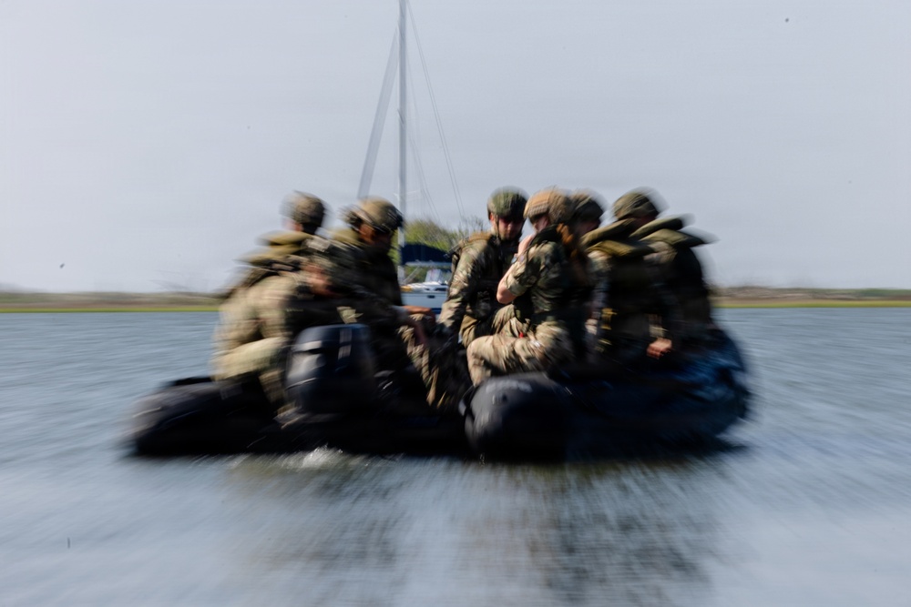 A group of military personnel in full tactical gear are traveling at high speed across a body of water in a dark inflatable rubber assault boat (RHIB/Zodiac-type). The image is intentionally motion-blurred, conveying a strong sense of speed and urgency. A sailboat mast and flat shoreline are visible in the background.