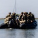 A group of military personnel in full tactical gear are traveling at high speed across a body of water in a dark inflatable rubber assault boat (RHIB/Zodiac-type). The image is intentionally motion-blurred, conveying a strong sense of speed and urgency. A sailboat mast and flat shoreline are visible in the background.