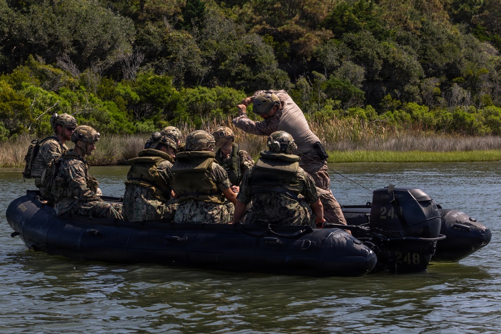 A group of military personnel in camouflage uniforms and tactical gear are aboard a dark inflatable rubber raiding craft (CRRC) on a calm waterway surrounded by marsh vegetation and trees. One individual standing at the bow appears to be giving directions or signals while others are seated and focused on a task. This appears to be a military training exercise, likely involving naval special warfare or amphibious operations.
