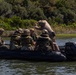 A group of military personnel in camouflage uniforms and tactical gear are aboard a dark inflatable rubber raiding craft (CRRC) on a calm waterway surrounded by marsh vegetation and trees. One individual standing at the bow appears to be giving directions or signals while others are seated and focused on a task. This appears to be a military training exercise, likely involving naval special warfare or amphibious operations.