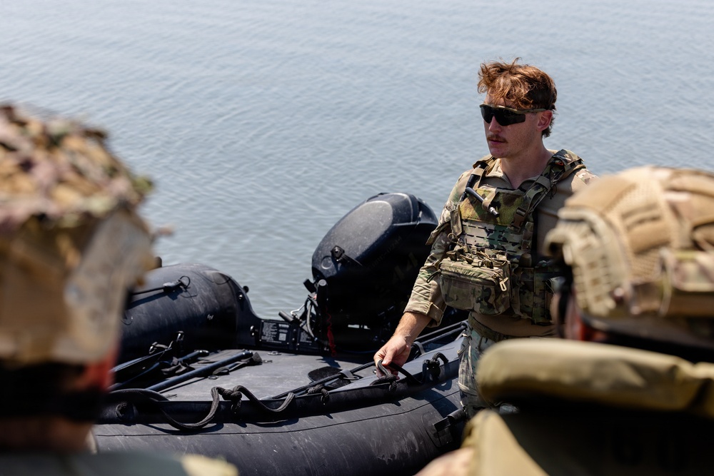 A military personnel wearing multicam camouflage uniform and plate carrier vest is standing near an inflatable rubber assault boat (CRRC or similar) on what appears to be a waterway. The soldier has reddish-brown hair, sunglasses, and a mustache, and appears to be securing or inspecting the watercraft. Additional personnel in similar gear are partially visible in the foreground.