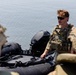 A military personnel wearing multicam camouflage uniform and plate carrier vest is standing near an inflatable rubber assault boat (CRRC or similar) on what appears to be a waterway. The soldier has reddish-brown hair, sunglasses, and a mustache, and appears to be securing or inspecting the watercraft. Additional personnel in similar gear are partially visible in the foreground.