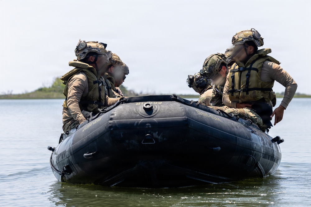 Four military personnel in full tactical gear are seated in a black inflatable combat rubber raiding craft (CRRC) on a calm body of water. The individuals are wearing camouflage uniforms, tactical vests, helmets with mounted accessories, and life preservers. Their faces have been digitally obscured, suggesting operational security considerations.
