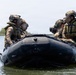 Four military personnel in full tactical gear are seated in a black inflatable combat rubber raiding craft (CRRC) on a calm body of water. The individuals are wearing camouflage uniforms, tactical vests, helmets with mounted accessories, and life preservers. Their faces have been digitally obscured, suggesting operational security considerations.
