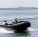 Two military personnel are operating a black rigid inflatable boat (RIB) at speed across open water. Both individuals are wearing military-style helmets and camouflage/tactical gear. A coastline with buildings and vegetation is visible in the background.