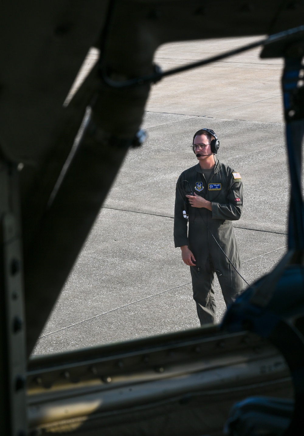 A military aviator or aircrew member in an olive green flight suit is standing on a concrete flight line, viewed through the open cargo door or hatch of a military aircraft. The individual is wearing a communications headset with a boom microphone and glasses, and appears to be monitoring or supervising ground operations. The photo is taken from inside the aircraft looking outward, creating a framed perspective.