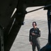 A military aviator or aircrew member in an olive green flight suit is standing on a concrete flight line, viewed through the open cargo door or hatch of a military aircraft. The individual is wearing a communications headset with a boom microphone and glasses, and appears to be monitoring or supervising ground operations. The photo is taken from inside the aircraft looking outward, creating a framed perspective.